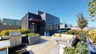 This rooftop terrace offers an expansive outdoor retreat elevated above a vibrant urban landscape. The space is defined by large-format, light-toned pavers that provide a clean, contemporary foundation throughout. A modern rooftop access structure clad in dark metal panel siding and horizontal louvers anchors one end of the terrace, lending an industrial-chic architectural character to the overall design.

The terrace is thoughtfully organized into distinct zones. A generous lounge area is furnished with plush wicker sectional seating accented by yellow and striped throw pillows, centered around a round coffee table—ideal for relaxed gatherings. Nearby, a teak dining table with cushioned chairs provides an al fresco dining option suited for both casual and formal occasions.

Raised planter beds, constructed from sleek concrete and filled with a variety of shrubs, flowering plants, and a deciduous tree, create natural privacy screens and introduce vibrant greenery throughout the space. A gravel garden border along the terrace perimeter adds textural contrast and further softens the hardscape.

Perimeter metal railings frame unobstructed panoramic views of the surrounding cityscape, including recognizable urban landmarks and hillside neighborhoods. The terrace enjoys abundant natural light and open sky, creating a bright and welcoming outdoor environment throughout the day.