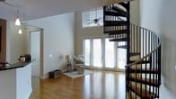 This well-designed living space makes an immediate impression with its soaring double-height ceilings that create an expansive, airy atmosphere. The room is anchored by warm hardwood flooring that extends throughout, adding a natural, cohesive quality to the space. A dramatic black metal spiral staircase serves as a striking architectural focal point, combining both form and function as it winds upward with dark wood treads that complement the flooring below.

Natural light floods the interior through a generous arrangement of floor-to-ceiling glass doors and large windows, including a clerestory window positioned high on the wall that draws the eye upward while enhancing the sense of vertical space. The glass doors open to the outside, offering easy access and views of the surrounding exterior.

The neutral, light-toned walls provide a clean backdrop, allowing the architectural elements to stand out. A ceiling fan is mounted overhead, adding practical comfort to the room. The living area is thoughtfully appointed with a cozy seating arrangement, a floor lamp, and a small area rug that defines the space. The overall ambiance is bright, modern, and inviting, with an open layout that feels both spacious and functional.