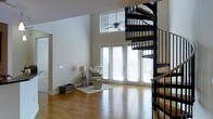 This well-designed living space makes an immediate impression with its soaring double-height ceilings that create an expansive, airy atmosphere. The room is anchored by warm hardwood flooring that extends throughout, adding a natural, cohesive quality to the space. A dramatic black metal spiral staircase serves as a striking architectural focal point, combining both form and function as it winds upward with dark wood treads that complement the flooring below.

Natural light floods the interior through a generous arrangement of floor-to-ceiling glass doors and large windows, including a clerestory window positioned high on the wall that draws the eye upward while enhancing the sense of vertical space. The glass doors open to the outside, offering easy access and views of the surrounding exterior.

The neutral, light-toned walls provide a clean backdrop, allowing the architectural elements to stand out. A ceiling fan is mounted overhead, adding practical comfort to the room. The living area is thoughtfully appointed with a cozy seating arrangement, a floor lamp, and a small area rug that defines the space. The overall ambiance is bright, modern, and inviting, with an open layout that feels both spacious and functional.