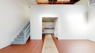 Located at 4401 San Leandro Street in Oakland, CA, this two-floor, one-bedroom, one-bathroom space offers a distinctive industrial character with raw architectural details throughout. The ground floor welcomes you with a generously open main area featuring terracotta-painted concrete floors, soaring ceilings with exposed wooden beams and corrugated metal roofing, and large windows that allow natural light to pour in. A staircase leads to the upper floor, while an open archway connects the main living area to the kitchen. The kitchen is well-appointed with white shaker-style cabinetry, granite-style countertops, a gas range, and a full-size refrigerator — all set beneath exposed beam ceilings that reinforce the space's industrial aesthetic. The upper floor continues the open feel with light gray painted floors, dramatically exposed roof framing with skylights, and ample open area suited for a variety of uses. The bathroom includes a full soaking tub with shower, built-in shelving, and a dark wood vanity with a stone-topped sink. White walls throughout create a clean, bright contrast to the raw structural elements, giving the space a compelling blend of utilitarian character and functional livability.