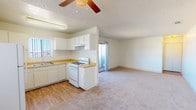 This single-story space offers a straightforward, functional layout where the kitchen and living area share an open, connected floor plan. The kitchen is equipped with white cabinetry, a gas range, a built-in sink, and laminate countertops in a warm wood tone. Wood-look flooring runs through the kitchen area, transitioning to plush carpet in the adjacent living space, creating a clear yet seamless division between the two zones. A textured ceiling adds dimension overhead, while a ceiling fan with a light fixture serves both as a practical comfort feature and a focal point above the living area. Natural light enters through a window near the kitchen sink. A sliding glass door visible in the background leads to an outdoor area, providing access to views of an expansive desert landscape with open terrain and distant structures on the horizon. The overall palette is neutral and light, with white walls and warm-toned flooring contributing to a clean, understated aesthetic. The open-concept design maximizes the sense of space throughout the interior.