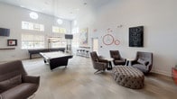 This single-floor amenity lounge is a well-appointed recreational and social space designed with a modern aesthetic and an eclectic artistic sensibility. The room commands immediate attention with its soaring double-height ceilings, which amplify the sense of openness and allow natural light to pour in through a series of large windows and a distinctive circular upper-level window. Recessed lighting and a sculptural pendant chandelier above the pool table provide warm, layered illumination throughout.

The space is anchored by a full-size pool table positioned centrally beneath the statement lighting fixture, while a shuffleboard table along the perimeter wall adds another recreational option. A floor-to-ceiling glass partition wall separates an adjacent lounge or workspace, maintaining visual connectivity throughout.

The lounge seating area features upholstered armchairs, a large tufted ottoman, and petite side tables arranged for comfortable conversation or relaxed viewing of the wall-mounted television. A media console with display storage and an orb-shaped table lamp add functional style to this corner.

Curated artwork and sculptural installations—including a wall-mounted penny-farthing bicycle, a hopscotch-themed wall panel, a figurative red sculpture, and large-format paintings—infuse the space with creative character. Light neutral flooring and white walls unify the design, creating an inviting, gallery-like atmosphere.