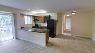 This single-floor space showcases an open-concept layout that seamlessly connects the kitchen and living areas. The kitchen is equipped with black appliances, including a full-size refrigerator, gas range with overhead exhaust, and a built-in microwave, all complemented by warm medium-toned wood cabinetry. A peninsula with a dark granite-style countertop serves as a natural divider between the kitchen and the adjoining living space, offering additional prep or seating potential. Recessed and flush-mounted ceiling lighting illuminate the kitchen area effectively. The living space features neutral wall-to-wall carpeting that extends into a hallway visible in the background, leading to additional rooms. The walls are painted in a soft, neutral tone throughout, contributing to a bright and cohesive ambiance. A window in the kitchen area allows natural light to filter in. The overall layout is practical and functional, with a straightforward flow between spaces making the unit feel open and easy to navigate.