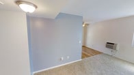 This single-floor space offers a clean, open layout designed for comfortable everyday living. A standout architectural detail is the vaulted ceiling, which adds a sense of height and airiness to the interior. The living area is defined by a soft, neutral carpet underfoot that transitions seamlessly into warm hardwood flooring in the adjoining space, creating a natural visual distinction between areas.

A prominent blue-gray accent wall serves as a focal point, adding a touch of color and character to the otherwise neutral palette of white walls and ceilings. The ceiling features a flush-mount light fixture in the carpeted area, while a ceiling fan with a light kit is installed in the hardwood-floored section, providing both air circulation and ambient lighting.

The walls are finished with a subtle texture and fitted with standard electrical outlets, reflecting a practical and functional design. Clean white baseboards frame the flooring throughout, adding a crisp, finished look. The overall ambiance of the space is bright, calm, and move-in ready, with a simple and versatile aesthetic that lends itself well to a variety of interior styles and furnishing choices.