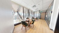 This contemporary commercial space is thoughtfully designed to serve as a collaborative lounge and workstation hub, spread across a single floor. The layout comprises two distinct zones that together create a cohesive, functional environment. The primary lounge area is defined by expansive floor-to-ceiling windows adorned with bold circular-patterned privacy film, allowing abundant natural light to filter in while framing impressive elevated city views. Light oak hardwood flooring runs throughout, complementing the warm tones of the round wooden bistro tables paired with ergonomic green-and-tan upholstered chairs arranged in intimate groupings. A steel-framed glass partition wall elegantly divides the lounge from the adjoining corridor, maintaining an open visual connection between spaces. The corridor area features a wall-mounted smart locker system in sleek grey, ideal for secure personal storage. Numbered private rooms line one side of the hallway, each accessed via bold black doors and complemented by vibrant botanical mural artwork along the walls. The drop ceiling with recessed tile panels and integrated HVAC vents ensures a clean, professional finish throughout. Together, the space balances aesthetic appeal with practical utility, making it well-suited for modern collaborative or hospitality use.