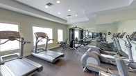 This fitness center offers a comprehensive workout environment designed to accommodate a range of exercise needs. The space is anchored by a dark, rubberized flooring that provides both durability and comfort during workouts. Multiple treadmills and an elliptical trainer are positioned near large windows, allowing natural light to fill the space and offering a bright, energizing atmosphere for cardio sessions. A multi-station cable machine serves as the centerpiece for strength training, complemented by additional resistance equipment arranged throughout the room. A full-length wall mirror spans one side of the facility, visually expanding the space and allowing users to monitor their form during exercise. The ceiling features a subtle tray detail with recessed lighting, contributing to a clean and polished aesthetic. HVAC vents are integrated into the ceiling for climate control, ensuring a comfortable environment year-round. The neutral gray and white color palette gives the space a contemporary, professional feel. Overall, the fitness center is thoughtfully laid out to maximize usability while maintaining an open, uncluttered atmosphere suitable for a variety of fitness routines.