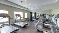 This fitness center offers a comprehensive workout environment designed to accommodate a range of exercise needs. The space is anchored by a dark, rubberized flooring that provides both durability and comfort during workouts. Multiple treadmills and an elliptical trainer are positioned near large windows, allowing natural light to fill the space and offering a bright, energizing atmosphere for cardio sessions. A multi-station cable machine serves as the centerpiece for strength training, complemented by additional resistance equipment arranged throughout the room. A full-length wall mirror spans one side of the facility, visually expanding the space and allowing users to monitor their form during exercise. The ceiling features a subtle tray detail with recessed lighting, contributing to a clean and polished aesthetic. HVAC vents are integrated into the ceiling for climate control, ensuring a comfortable environment year-round. The neutral gray and white color palette gives the space a contemporary, professional feel. Overall, the fitness center is thoughtfully laid out to maximize usability while maintaining an open, uncluttered atmosphere suitable for a variety of fitness routines.