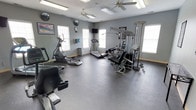 This fitness center offers a comprehensive workout environment designed to accommodate a range of exercise needs. The space is outfitted with durable rubber flooring, providing a safe and practical surface for both cardio and strength training activities. Multiple windows line the walls, flooding the room with natural light and creating an open, energizing atmosphere. Ceiling fans are positioned throughout to maintain comfortable airflow during workouts.

The cardio section features a treadmill, an elliptical trainer, and a stationary bike, offering users a variety of low-impact and aerobic exercise options. For strength training, the space includes a multi-station cable machine and a weight bench with attachments, enabling a full range of resistance exercises targeting different muscle groups. A wall-mounted television is installed for entertainment, while a wall clock and a fitness reference chart add functional touches to the space.

The overall ambiance is clean, practical, and well-maintained, with light gray walls contributing to a neutral and focused environment. The combination of natural light, ventilation, and a diverse selection of equipment makes this fitness center a functional and inviting space for everyday training.
