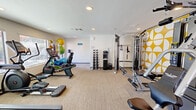 Located at 7425 N Mona Lisa Rd in Tucson, Arizona, this fitness center offers a thoughtfully organized exercise space designed to support a wide range of workout routines. The room is finished with neutral carpeting and crisp white walls, creating a clean and energizing environment. A bold, geometric yellow-and-white accent wall serves as a striking focal point, adding a modern and vibrant character to the space.

The fitness center is outfitted with a comprehensive selection of equipment. Cardio options include treadmills, a stationary recumbent bike, and an elliptical machine, all positioned to take advantage of natural light from large windows that offer views of the outdoor pool area. A multi-station cable machine and a weight bench provide dedicated strength training capabilities, while a tiered dumbbell rack with a range of free weights supports varied resistance training.

A functional training corner houses resistance bands, stability balls in multiple sizes, yoga mats, and balance accessories, making the space adaptable for stretching, group exercise, and dynamic training. Two wall-mounted flat-screen televisions are positioned for easy viewing during cardio sessions. Recessed lighting throughout ensures consistent illumination across the entire floor. The overall layout is efficient and user-friendly, catering to both casual exercisers and dedicated fitness enthusiasts.