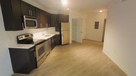 This single-floor unit showcases an open-concept layout that seamlessly connects the kitchen and living areas, creating a spacious and functional environment. The kitchen is well-appointed with dark espresso-toned cabinetry that contrasts sharply against light countertops, lending a modern, contemporary aesthetic. Stainless steel appliances, including a refrigerator, range, dishwasher, and over-the-range microwave, provide a sleek and cohesive look. A double-basin sink is positioned beneath upper cabinetry, maximizing storage and workspace efficiency. Track lighting above the kitchen area ensures ample illumination for food preparation. Light hardwood-style flooring runs throughout the open space, contributing to a bright and airy atmosphere. The living area extends naturally from the kitchen, offering flexible space for various furniture arrangements. A utility panel is visible along one wall, and a doorway leads to additional rooms beyond, suggesting further private spaces within the unit. The overall design balances practicality with contemporary style, making it a functional and visually cohesive living space.