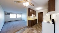 This single-floor space features an open-concept layout that connects the kitchen and living area, creating a functional and cohesive floor plan. The kitchen is equipped with dark wood cabinetry both above and below the counter, a single-basin sink, light-colored laminate countertops, a white refrigerator, and a white electric range. The flooring in the kitchen area consists of a durable tile with a classic diamond pattern in earthy tones. A closet with sliding wood-paneled doors is conveniently situated just off the kitchen, offering additional storage. The living area transitions seamlessly from the kitchen, featuring plush carpeting and a wide window that allows natural light to fill the room. A baseboard heater runs along the wall beneath the window, providing supplemental warmth. A ceiling fan with a light fixture is centrally positioned, serving both the kitchen and living areas. The walls and ceilings are neutral in tone, with a textured ceiling finish throughout. The overall design reflects a practical, no-frills layout well-suited for everyday living.