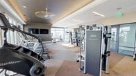 This contemporary fitness center is thoughtfully designed to provide a comprehensive workout environment. The space features a striking combination of light and dark architectural elements, with a raised tray ceiling accented by warm ambient lighting and ceiling fans that contribute to a comfortable training atmosphere. Large floor-to-ceiling windows line the perimeter, flooding the space with natural light and offering open outdoor views that enhance the overall sense of spaciousness.

The cardio zone is outfitted with a row of Star Trac treadmills, elliptical trainers, and stair climbers, all positioned to take advantage of the expansive window views. Mounted flat-screen televisions provide entertainment throughout the space. The strength training area features a range of Matrix Fitness and Nautilus Instinct cable and selectorized machines, along with free-weight stations equipped with benches and barbells.

A dedicated free weights section houses a full rack of dumbbells and kettlebells, neatly organized along mirrored walls that visually expand the area. Built-in cabinetry with stone countertops adds a refined, upscale touch to the free weights zone. Geometric patterned carpet tiles in neutral tones of grey and warm brown unify the space while defining distinct workout zones. The overall ambiance is polished, modern, and purpose-built for serious fitness.