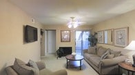 This single-floor space is designed for comfortable everyday living, centered around a welcoming living room that serves as the heart of the home. The living room is finished with warm, neutral-toned walls and soft carpeted flooring, creating a cozy and inviting atmosphere. A ceiling fan with an integrated light fixture anchors the room from above, providing both airflow and ambient lighting. Natural light floods the space through a large sliding glass door dressed with vertical blinds, which opens onto a balcony overlooking a lush outdoor area with mature greenery. The room comfortably accommodates generous seating, a round wood coffee table, and a wall-mounted flat-screen television, making it well-suited for both relaxation and entertaining. Built-in shelving is visible along one wall, offering practical storage and display space. Decorative framed artwork adds character to the walls, while potted plants bring a touch of natural life indoors. A hallway visible from the living area leads to additional rooms, suggesting a thoughtfully laid-out floor plan. Overall, the space strikes a balance between functionality and warmth, with a light, airy feel enhanced by its connection to the outdoors.