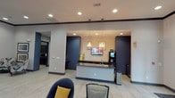 This single-floor commercial space presents a sleek, contemporary design centered around a functional island-style counter with a dark countertop and built-in sink. The counter serves as a focal point, flanked by dark navy accent columns and walls that contrast with the light neutral tones of the tile flooring. Overhead, a combination of recessed lighting and elegant pendant fixtures with exposed bulbs provides warm, layered illumination throughout the space.

A textured accent wall behind the counter adds visual depth, while a wall-mounted flat-screen television and a beverage station contribute to the space's utility and comfort. The ceiling features subtle architectural detailing, including a defined border trim that frames the central area.

The seating area in the foreground includes upholstered chairs in deep blue tones, accented with a mustard yellow throw pillow, offering a welcoming and stylish area for occupants. Adjacent areas visible through open passages suggest additional functional rooms, including what appears to be a kitchenette or service area fitted with dark cabinetry.

Overall, the space balances functionality with modern aesthetic appeal, making it well-suited for use as a hospitality lobby, corporate lounge, or community gathering area.