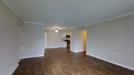 This single-floor residence showcases an open-concept layout that seamlessly connects the main living area to the kitchen. The living space is finished with rich, dark hardwood-style flooring that runs throughout, providing a warm and cohesive aesthetic. Crown molding lines the ceilings, adding a refined architectural detail to the otherwise clean, neutral walls painted in a soft gray-white tone.

The kitchen is equipped with stainless steel appliances, including a range, microwave, and refrigerator, complemented by a decorative tile backsplash that adds a subtle design accent. White cabinetry provides ample storage and contributes to a bright, functional cooking space. A ceiling fan is centrally positioned above, ensuring comfortable airflow throughout the open area.

A doorway visible to the left suggests access to additional rooms, while a hallway on the right leads further into the unit. The overall ambiance is clean, modern, and move-in ready, with consistent finishes throughout that create a unified and practical living environment suited for everyday comfort.