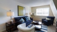 This single-floor residence offers a comfortable and thoughtfully arranged living space designed for everyday functionality. The living room serves as the centerpiece of the home, featuring a large navy blue sofa adorned with decorative throw pillows in coordinating blues and greens, complemented by a sleek white accent chair and a round, dark-toned coffee table. A striped area rug anchors the seating arrangement, adding a layer of visual interest and texture to the room. A bold, yellow-toned framed artwork displayed on the wall provides a striking focal point, while a pair of table lamps contribute to the warm, layered lighting throughout the space. Natural light filters generously through a large window fitted with horizontal blinds, brightening the neutral-toned walls and lending an airy quality to the interior. A baseboard heating unit runs along the lower wall beneath the window, highlighting the practical amenities built into the space. The overall ambiance is inviting and well-balanced, blending casual comfort with clean, simple design elements that create a livable and approachable atmosphere throughout.