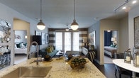 This single-story Dallas residence showcases a well-designed open-concept floor plan that connects the kitchen, living area, and bedroom in a cohesive, functional layout. The kitchen serves as a central focal point, featuring a substantial granite-topped island with warm gold and cream veining, complemented by a stainless steel sink with a high-arc faucet. Two globe pendant lights suspended from chains hang above the island, providing both task lighting and a stylish visual anchor.

The living area is defined by dark hardwood flooring and a neutral gray color palette, creating a sophisticated backdrop for the furnished space. A teal sofa with decorative accent pillows anchors the seating area, positioned in front of French doors that allow natural light to filter through striped horizontal curtains. A ceiling fan centered between the doors enhances the room's functionality.

Framed artwork on the gray walls adds visual interest, while a bedroom is partially visible through a doorway at the far right, furnished with navy blue drapery that contributes to the overall cohesive color scheme. The interplay of warm and cool tones throughout creates a balanced, inviting ambiance.