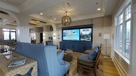 Located at 722 Island Park Drive on Daniel Island, South Carolina, this single-story space exudes a coastal, resort-inspired aesthetic that blends comfort with refined design. The lounge area serves as a central gathering point, anchored by a large built-in media console finished in deep navy blue, housing an expansive flat-screen display that commands attention. Crown molding and wainscoting detail the walls, adding architectural character throughout. A decorative orb chandelier crafted from natural materials hangs overhead, complementing the warm hardwood-style flooring beneath. Recessed lighting and wall sconces provide layered illumination, contributing to an inviting ambiance. Comfortable seating, including upholstered armchairs and sofas in coordinating blue tones with patterned accent pillows, is arranged to encourage conversation and relaxation. A gently arched doorway with white pilasters frames the transition into an adjacent area, hinting at the continuity of thoughtful design throughout the space. The overall atmosphere balances casual coastal charm with polished, community-oriented functionality.
