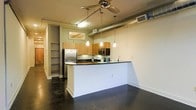 This single-floor space showcases a thoughtfully designed open-concept layout that seamlessly integrates the kitchen and living areas. The kitchen is a standout feature, equipped with stainless steel appliances, including a full-size refrigerator and built-in microwave, complemented by warm wood cabinetry and dark countertops. A wraparound peninsula provides additional workspace and doubles as a casual dining or entertaining surface.

The industrial character of the space is evident in the exposed metallic ductwork running along the ceiling, which adds an urban, loft-inspired aesthetic. Pendant lights hang above the peninsula, providing focused task lighting, while a ceiling fan ensures comfortable airflow throughout the open area. The polished dark concrete flooring grounds the space with a sleek, low-maintenance surface that reinforces the contemporary industrial feel.

A hallway visible from the main living area leads to additional rooms and features built-in shelving, offering practical storage solutions. A small transom-style window allows natural light to filter into the interior corridor. Overall, the space balances functional design with modern urban aesthetics, making it a versatile and stylish environment.