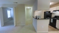 This single-floor residence offers a clean and functional layout highlighted by neutral, light-toned walls and soft carpeting that runs throughout the main living areas and hallway. The space is designed with practicality in mind, featuring a well-positioned kitchen area with a raised breakfast bar countertop finished in dark stone, providing a natural division between the kitchen and the adjacent living space. White cabinetry lines the kitchen walls, offering ample storage, while a black refrigerator anchors the appliance suite. The hallway extends the length of the unit, leading to additional rooms and creating a clear, organized flow through the space. Overhead lighting provides consistent illumination along the corridor, complementing the warm, understated color palette. The trim and baseboards throughout are neatly finished in white, adding a crisp contrast to the beige walls. The overall ambiance is calm and neutral, offering a versatile backdrop well-suited for a variety of interior styles. The single-story layout ensures ease of accessibility and seamless movement between all areas of the home.