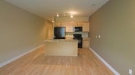 This single-floor space features an open-concept layout that seamlessly integrates the kitchen and living areas, creating a spacious and functional environment. The light hardwood floors extend throughout, contributing to a bright, cohesive aesthetic that enhances the sense of openness.

The kitchen is well-appointed with light maple cabinetry providing ample storage. A peninsula with granite countertops serves as both a prep area and an informal dining or gathering space, with a built-in sink adding practicality. Black appliances, including a refrigerator, electric range, and over-the-range microwave, offer a modern contrast to the lighter tones of the cabinetry and countertops.

Track lighting runs across the kitchen ceiling, ensuring the workspace is well-lit. The walls are finished in a warm neutral tone that complements the natural wood flooring and cabinetry throughout. White baseboards provide clean contrast along the perimeter.

The adjacent open living area benefits from the natural flow of the space, offering flexibility for various furniture arrangements. The overall ambiance is clean, modern, and inviting, making the space highly functional for everyday living.
