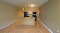 This single-floor space features an open-concept layout that seamlessly integrates the kitchen and living areas, creating a spacious and functional environment. The light hardwood floors extend throughout, contributing to a bright, cohesive aesthetic that enhances the sense of openness.

The kitchen is well-appointed with light maple cabinetry providing ample storage. A peninsula with granite countertops serves as both a prep area and an informal dining or gathering space, with a built-in sink adding practicality. Black appliances, including a refrigerator, electric range, and over-the-range microwave, offer a modern contrast to the lighter tones of the cabinetry and countertops.

Track lighting runs across the kitchen ceiling, ensuring the workspace is well-lit. The walls are finished in a warm neutral tone that complements the natural wood flooring and cabinetry throughout. White baseboards provide clean contrast along the perimeter.

The adjacent open living area benefits from the natural flow of the space, offering flexibility for various furniture arrangements. The overall ambiance is clean, modern, and inviting, making the space highly functional for everyday living.