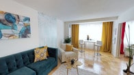 This thoughtfully designed single-floor space showcases a cohesive and vibrant interior aesthetic anchored by a rich teal velvet sofa and warm golden-yellow accents throughout. The living area serves as the focal point, featuring a large-scale abstract wall panel in soft aqua tones that adds an artistic, gallery-like quality to the space. A plush cream armchair and a sculptural gold coffee table complement the seating arrangement, while a textured white area rug grounds the layout with understated elegance. The flooring consists of classic parquet wood tiles, lending warmth and timeless character across the open layout. Decorative elements such as patterned throw pillows, a globe ornament, and a potted plant introduce layers of texture and life to the room. Toward the front of the space, a bright dining nook is framed by floor-length golden curtains that frame a generous window, flooding the area with natural light. A sleek white bar-height table paired with modern white chairs offers a clean, contemporary contrast to the warmer tones of the living zone. The overall ambiance strikes a balance between bold and inviting, with a carefully curated color palette and a mix of modern furnishings that give the space a polished yet comfortable feel.