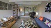 This single-floor residence offers a thoughtfully designed open-concept layout that seamlessly connects the living, dining, and entryway areas into one fluid, functional space. The living area is anchored by a plush navy sofa, a geometric glass-top coffee table resting on a bold chevron rug, and an industrial-style wood and metal shelving unit that adds both storage and character. Hardwood-style flooring flows consistently throughout, lending warmth and visual continuity to the entire space. Structural beams accent the ceiling, adding an architectural dimension that enhances the room's character. Moving toward the dining area, a mid-century modern table and chairs sit beneath a ceiling fan with a light fixture, creating a casual yet refined dining experience. A leather accent chair with a side table and lamp occupies a transitional nook, offering a cozy reading or relaxation spot. The entryway beyond features a coat rack and closet, providing practical organization near the entrance. Neutral gray walls serve as a versatile backdrop, allowing the furnishings and decor to take center stage. The overall ambiance is modern and inviting, blending industrial and mid-century influences for a space that feels both curated and comfortable.
