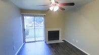 This single-story space presents a clean, functional layout anchored by a well-appointed living area. The room features dark hardwood-style flooring that runs throughout, providing a warm and polished foundation that contrasts attractively with the neutral beige walls. A central fireplace with a light-toned surround and raised hearth serves as a natural focal point, adding both character and a cozy ambiance to the space.

Natural light flows generously into the room through a full-width sliding glass door fitted with vertical blinds, offering views of a lush outdoor area with mature trees visible beyond. This door also provides convenient access to an outdoor balcony or patio, blending the interior and exterior environments seamlessly.

Overhead, a ceiling fan with an integrated light fixture provides both ventilation and ambient lighting, contributing to the room's practical and comfortable atmosphere. The clean, uncluttered walls and simple trim work give the space a modest, move-in-ready quality. Overall, the living area balances everyday functionality with inviting details, making it a versatile and comfortable environment.
