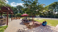 Located in Huntsville, Alabama, this thoughtfully designed outdoor amenity space offers a welcoming environment for recreation and relaxation. The grounds are anchored by a well-maintained brick paver courtyard that serves as the central gathering hub, furnished with picnic tables shaded by vibrant red umbrellas and a wooden pergola structure that adds architectural character to the setting. Adirondack-style seating is arranged throughout, creating comfortable spots for casual conversation and leisure. Mature crape myrtles and large shade trees are strategically placed across the landscape, providing natural canopy and colorful seasonal blooms that enhance the overall ambiance. Decorative planters with bold blue glazing add distinct visual interest throughout the space. A dedicated tennis court sits adjacent to the main courtyard area, enclosed by chain-link fencing and surrounded by manicured lawn. A cornhole area with a sand pit offers additional recreational options. Curved pathways of both pavers and poured concrete connect the various activity zones, guiding visitors seamlessly through the grounds. The exterior building facade features a clean, vertical white panel design complemented by neatly trimmed foundation plantings. The overall setting is open, airy, and well-lit by natural sunlight, making it an inviting outdoor destination throughout the warmer months.
