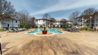 Located at 6303 Delaney Dr in Kannapolis, North Carolina, this outdoor amenity space centers around a generously sized resort-style swimming pool with clear, blue-green water. The pool features a curved, freeform design with multiple entry points via stainless steel handrails, and is surrounded by an expansive stamped concrete deck that provides ample space for relaxation and socializing. Decorative large planters with seasonal greenery serve as elegant focal points throughout the deck area.

Multiple pergola structures are positioned around the perimeter, offering shaded retreats and framing the space with architectural character. An array of lounge chairs and seating areas are thoughtfully arranged poolside, catering to both sun-seekers and those preferring shade. A distinct second pool area is visible, indicating a dual-pool setup for varied recreational use.

The surrounding landscaping features manicured topiary shrubs, ornamental grasses, and mature trees, contributing to a lush, well-maintained environment. Curved stone and brick retaining walls with stamped paver pathways add refined hardscape detailing throughout the grounds. The community buildings visible in the background feature multi-story exteriors with balconies, complementing the upscale outdoor setting. Overall, the space delivers a polished, resort-inspired atmosphere ideal for leisure and outdoor living.