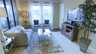 This single-story space showcases a thoughtfully designed living area that balances classic elegance with warm, rustic touches. The living room is anchored by two tall windows dressed in sheer white curtains, allowing an abundance of natural light to fill the room while offering views of an outdoor balcony railing and surrounding greenery. The neutral, warm wall tones create a welcoming backdrop for the carefully curated furnishings.

The seating arrangement features a plush gray sofa paired with two ornate, French-style accent chairs upholstered in a soft blue-gray fabric with silver-toned carved wood frames, adding a refined, vintage-inspired character to the space. A sleek glass-top coffee table with a metallic frame sits atop a generous, textured shag area rug, grounding the seating area with layers of comfort and visual interest.

On the opposite wall, a rustic reclaimed-wood media console provides a striking contrast to the softer furnishings, supporting a large flat-screen television. A coordinating side table with a decorative lamp and floral arrangement completes the room, contributing to the space's polished yet inviting ambiance.
