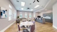 Located at 5095 Big Lake Rd in Lake Charles, Louisiana, this single-floor communal space offers a thoughtfully designed open-plan layout that seamlessly integrates a living room, dining area, and kitchen. Warm, honey-toned luxury vinyl plank flooring runs throughout, complementing the soft lavender-gray walls and bright white trim for a clean, contemporary aesthetic.

The kitchen is outfitted with gray shaker-style cabinetry extending from floor to ceiling, granite-look countertops, a stainless steel sink, and black appliances including a full-size refrigerator and microwave. A central island with bar seating provides an ideal space for casual dining or gathering.

The dining area features a round white table with upholstered purple accent chairs, adding a pop of color to the neutral palette. The living room is generously furnished with gray sofas, armchairs, and layered area rugs, creating an inviting lounge atmosphere. Large windows with white blinds line multiple walls, flooding the space with natural light and offering views of the surrounding greenery.

Elevated ceilings enhance the sense of openness, while recessed lighting ensures a well-lit environment. The space also features glass-paneled interior doors and clearly marked exits, indicating its suitability for a community or facility setting.