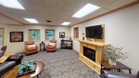 This inviting single-floor common space is thoughtfully designed to accommodate a variety of social activities and comfortable gatherings. The layout flows naturally between distinct functional zones, each finished with warm honey-toned wood accents, a drop tile ceiling with recessed panel lighting, and light tile flooring throughout the main areas.

A prominent wet bar runs along one wall, featuring an extended wood-trimmed counter with a dark stone surface and a row of cushioned bar stools. A series of framed artwork lines the wall above, and a set of sliding glass doors at the end of the bar area opens directly to an outdoor patio, allowing natural light to fill the space and providing easy indoor-outdoor access.

Adjacent to the bar, a casual dining area offers small tables and chairs, creating an informal setting for conversation or card games. A compact kitchenette sits nearby, equipped with wood cabinetry, a stainless steel refrigerator, and a sink — well-suited for light food and beverage preparation.

The lounge area features patterned carpeting and is anchored by a wood-framed electric fireplace topped with a wall-mounted flat-screen television. Comfortable seating, including upholstered armchairs and a leather sofa, is arranged to encourage relaxed conversation. Windows along one wall bring in views of surrounding greenery, adding a serene, natural element to the overall ambiance.