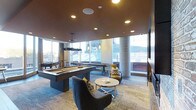 Located at 3737 Chestnut Street in Philadelphia, PA, this single-floor amenity lounge is designed for both relaxation and recreation. The space showcases a modern, upscale aesthetic defined by a warm, rich ceiling finish that contrasts beautifully with the light-toned patterned carpeting below. Expansive floor-to-ceiling windows line the perimeter, allowing abundant natural light to flood the interior while offering views of the surrounding urban environment. Prominent concrete columns add an industrial architectural element that complements the contemporary design. At the heart of the space is a sleek, dark-finished pool table accompanied by stylish pendant lighting overhead. Adjacent to it, a shuffleboard table provides additional entertainment options. The lounge area features plush curved accent chairs, patterned throw pillows, and a polished round coffee table, creating an inviting gathering spot. A large flat-screen television is mounted on the wall, enhancing the entertainment experience. Recessed ceiling lighting supplements the natural light, ensuring the space remains well-lit throughout. The overall ambiance strikes a balance between sophisticated design and casual comfort, making it a well-appointed communal destination.