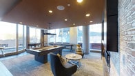 Located at 3737 Chestnut Street in Philadelphia, PA, this single-floor amenity lounge is designed for both relaxation and recreation. The space showcases a modern, upscale aesthetic defined by a warm, rich ceiling finish that contrasts beautifully with the light-toned patterned carpeting below. Expansive floor-to-ceiling windows line the perimeter, allowing abundant natural light to flood the interior while offering views of the surrounding urban environment. Prominent concrete columns add an industrial architectural element that complements the contemporary design. At the heart of the space is a sleek, dark-finished pool table accompanied by stylish pendant lighting overhead. Adjacent to it, a shuffleboard table provides additional entertainment options. The lounge area features plush curved accent chairs, patterned throw pillows, and a polished round coffee table, creating an inviting gathering spot. A large flat-screen television is mounted on the wall, enhancing the entertainment experience. Recessed ceiling lighting supplements the natural light, ensuring the space remains well-lit throughout. The overall ambiance strikes a balance between sophisticated design and casual comfort, making it a well-appointed communal destination.
