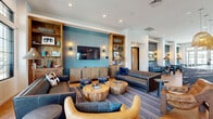 Located at 6250 S 40th St in Phoenix, AZ, this thoughtfully designed single-floor amenity space offers a generous layout spanning a substantial footprint. The entry welcomes visitors with herringbone hardwood flooring, soaring ceilings, floor-to-ceiling black-framed windows, and curated greenery set against a striking teal accent wall.

The living room serves as the heart of the space, featuring rich leather sofas, sculptural wood coffee tables, and built-in wood shelving flanking a wall-mounted flat-screen TV. Teal accent walls, woven pendant lighting, and striped area rugs lend a warm yet contemporary atmosphere. Large windows flood the seating areas with natural light and offer views of the surrounding Phoenix landscape.

A secondary lounge nook provides an intimate retreat, furnished with modular sectional sofas in navy and gray, speckled patterned carpeting, gold-accented side tables, and oversized framed artwork that enhances the curated aesthetic.

The dining area flows seamlessly from the lounge, anchored by a dramatic dark marble island, pendant lighting, and additional seating that connects to the broader communal space.

A dedicated conference room completes the layout, equipped with a sleek wood conference table, ergonomic seating, a wall-mounted display, built-in cabinetry, and bold teal walls accented by a statement art piece — ideal for productive meetings.