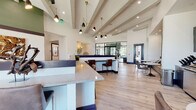 This single-floor communal space showcases a sophisticated contemporary design anchored by dramatic exposed beam ceilings that soar overhead, creating an airy, expansive atmosphere throughout. Light hardwood flooring runs continuously across the open-plan layout, unifying the various functional zones seamlessly.

The main gathering area features a generously sized lounge with a bold mustard leather sectional sofa, wicker accent chairs, and curated décor including sculptural pendants and gallery-style artwork. Adjacent to this, a central counter with a stone-topped surface serves as a reception or bar area, complemented by bar-height seating.

Built-in booth-style banquette seating with upholstered cushions and decorative geometric wall panels defines a secondary lounge zone, while a dedicated dining nook near floor-to-ceiling black-framed windows offers views of palm-lined outdoor surroundings, flooding the space with natural light.

Black-framed glass partition walls divide a private room or conference area, maintaining visual openness while providing separation. Recessed lighting and sculptural multi-arm pendant fixtures provide layered illumination throughout. The overall ambiance is modern, welcoming, and thoughtfully designed for both social interaction and relaxed gathering.