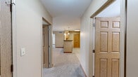 This well-appointed two-bedroom, two-bathroom residence at 1410 Deer Lakes Dr in Buffalo, NY offers a comfortable and functional single-floor layout. The open-concept kitchen and living area serves as the heart of the home, featuring warm honey-toned cabinetry, black appliances including a refrigerator, range, and dishwasher, light countertops, and pendant lighting over the peninsula. Light tile-style flooring runs throughout the kitchen, while plush neutral carpeting extends into the living and bedroom areas. The living room is bright and welcoming, with large windows fitted with white blinds that invite natural light, along with a private entry door. Both bedrooms are generously proportioned, with the primary suite offering a ceiling fan, windows with outdoor views, and access to a walk-in closet with wire shelving. The two bathrooms differ in configuration: one features a full tub/shower combination with wood-toned vanity cabinetry and tile flooring, while the second is designed for accessibility, offering a roll-in shower, pedestal sink, grab bars, and easy-access layout. A dedicated in-unit laundry room houses a stacked washer and dryer alongside a utility countertop. Rich wood-panel doors and warm neutral wall tones contribute to a cohesive, inviting ambiance throughout.