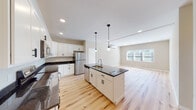 This thoughtfully designed single-floor unit blends modern finishes with a clean, contemporary aesthetic throughout. The heart of the space is an open-concept kitchen and living area anchored by a large central island with dark granite countertops, an undermount sink, and stylish pendant lighting above. White shaker-style cabinetry lines the kitchen walls, complemented by stainless steel appliances including a refrigerator, range, and built-in microwave. Light-toned hardwood floors with natural variation flow seamlessly through the kitchen, living, and bedroom areas, creating a warm and cohesive feel. The living area is bright and open, benefiting from natural light through a wide window. The bedroom is generously proportioned, featuring a double-door closet with matte black hardware, a wall-mounted mini-split climate control unit, and additional natural light from an exterior-facing window. Two bathrooms serve the unit — one full bath with a tiled tub-shower combo and dark granite vanity, and a second bath with a sleek white vanity, dark granite countertop, framed mirror, and elegant marble-look tile flooring. Matte black hardware and fixtures appear as consistent design accents throughout, lending the space a cohesive, modern character.