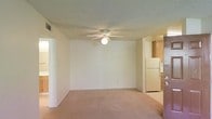 This single-floor residential space offers a clean, functional layout designed for everyday comfort. The main living area is finished with wall-to-wall carpeting in a neutral tone, creating a warm and understated atmosphere. A ceiling fan with an integrated light fixture is centered overhead on a textured ceiling, providing both air circulation and ambient lighting to the room. The walls are painted in a soft, neutral hue that contributes to a bright and open feel throughout the space. An HVAC vent is visible near the ceiling, indicating a climate control system integrated into the unit. Adjacent to the living area, the kitchen is accessible via an open passthrough, featuring warm wood-toned cabinetry and a white refrigerator, offering practical storage and appliance space. A secondary doorway on the opposite side of the living area suggests access to additional rooms or a hallway, hinting at a connected, flowing floor plan. Overall, the space presents a straightforward residential environment with a neutral palette, practical finishes, and a layout that emphasizes ease of use and comfortable day-to-day living.