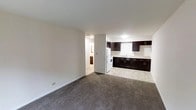 This single-floor unit features an open-concept design that seamlessly connects the living area to the kitchen. The living space is finished with wall-to-wall grey carpeting, creating a comfortable and cohesive atmosphere. Clean white walls and a smooth white ceiling keep the space feeling bright and open.

The kitchen is outfitted with dark espresso-toned cabinetry, both upper and lower, providing a strong visual contrast against the white walls and light-toned tile flooring. A white refrigerator and white range anchor the kitchen, accompanied by a single-basin sink set into a light countertop. The cabinetry offers ample storage, with upper cabinets extending toward the ceiling.

A hallway visible from the main living area leads to additional rooms, including what appears to be a bathroom, providing a functional and straightforward layout. The overall design is practical and clean, offering a neutral palette that accommodates a variety of interior styles. The unit presents a well-maintained space suitable for everyday living.