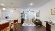 This single-floor residence is designed with a clean, modern aesthetic and a functional open-concept layout. Dark hardwood flooring runs consistently throughout the space, providing a warm contrast to the bright white walls and ceilings equipped with ceiling fans.

The living area is spacious and welcoming, with large windows dressed in sheer curtains that allow ample natural light to fill the room. A dedicated nook within the living space offers a quiet area suitable for a home office or reading corner.

The kitchen is a standout feature, showcasing white shaker-style cabinetry extending to the ceiling, granite countertops, a subway tile backsplash, and a full suite of stainless steel appliances including a refrigerator with an ice maker, range, microwave, and dishwasher. A large center island with seating for two serves as both a prep space and casual dining area, illuminated by pendant lighting above.

The bedroom is generously sized, featuring a ceiling fan, large windows with natural light, and ample wall space. The bathroom is elongated and well-appointed, with a granite-topped double vanity with white cabinetry, a soaking tub with a tiled surround, and dark hardwood flooring continuing throughout.