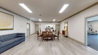 This single-story community space in Martinez, California is thoughtfully designed to accommodate gathering, dining, and relaxation across several interconnected zones. The layout flows openly between areas, unified by light wood-tone luxury vinyl plank flooring and neutral white walls that create a clean, welcoming atmosphere throughout.

The dining area features substantial rustic solid-wood tables paired with matching wood chairs and benches, offering generous seating capacity for group meals or events. Overhead, recessed panel lighting provides consistent, even illumination across the space.

An integrated kitchen is outfitted with white shaker-style cabinetry, grey quartz countertops, stainless steel appliances including a range, over-the-range microwave, and dishwasher. A central island with a bar stool provides additional workspace and casual seating.

Adjacent lounge areas offer distinct seating arrangements. One zone features two upholstered accent chairs with gold frames alongside a grey sofa, accented by framed artwork and a floor lamp. A second lounge area presents a pair of white barrel chairs, a gold accent table, a textured area rug, and decorative shelving, creating a more intimate setting. The overall ambiance is modern, clean, and functional.