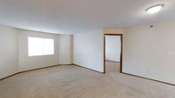 This single-floor space presents a straightforward and functional layout highlighted by a clean, neutral aesthetic throughout. The main living area is finished with wall-to-wall beige carpeting that adds warmth underfoot, complemented by crisp white walls and wood-toned baseboards that frame the room with subtle contrast. A large window along one wall allows natural light to fill the space, creating a bright and open atmosphere. The textured white ceiling adds a modest architectural detail overhead. An open doorway with a warm wood-toned trim connects the main living area to an adjoining room, suggesting a seamless flow between spaces. The adjoining room mirrors the same neutral palette and carpeted flooring, maintaining a cohesive look throughout the unit. The overall design is understated and versatile, providing a blank canvas well-suited to a variety of furnishing styles. The absence of clutter and the consistent use of light, neutral finishes throughout contribute to an airy, open feel across the entire floor.