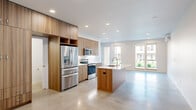 This contemporary two-story home offers five bedrooms and four bathrooms across a thoughtfully designed layout. The ground floor features polished concrete flooring throughout, beginning with a clean, minimalist entryway that opens into a bright, open-concept kitchen and living area. The kitchen is outfitted with warm wood-grain cabinetry, a white quartz waterfall island with an integrated sink, stainless steel appliances including a French door refrigerator, and a classic subway tile backsplash. Recessed lighting and large windows allow natural light to fill the space. The ground floor also includes a ground-level bedroom with polished concrete floors, a wall-mounted mini-split unit, and windows overlooking a green yard, as well as an attached garage with direct interior access. The upper floor transitions to light hardwood flooring and hosts the remaining bedrooms, including a generously sized primary suite featuring a dramatic skylight that floods the room with natural light and an en-suite bathroom. Additional bedrooms on this floor offer sliding glass doors with neighborhood views and sliding-door closets with built-in shelving. Throughout the home, crisp white walls, clean lines, and modern finishes create a cohesive, contemporary aesthetic.