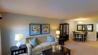 This single-floor residence offers a thoughtfully designed living space that balances comfort and style. The living room serves as the heart of the home, centered around a plush, cream-colored sofa accented with light blue throw pillows that add a pop of color against the soft, neutral-toned walls. A dark wood coffee table sits at the forefront, decorated with simple, elegant accents that enhance the room's welcoming ambiance. Two framed geometric art pieces hang side by side on the main wall, providing a modern yet understated focal point. Warm, layered lighting from table lamps creates a cozy and inviting atmosphere throughout. Transitioning from the living area, a dark wood china cabinet with glass-front doors adds a touch of classic elegance, serving as a natural divider between the living and dining spaces. The dining area features dark wood chairs arranged neatly, complementing the rich tones of the cabinetry. Neutral-toned carpeting flows throughout the space, tying the rooms together cohesively. The overall ambiance is calm and refined, making this space well-suited for both relaxed everyday living and comfortable entertaining.