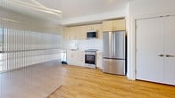 Located in South Salt Lake, Utah, this thoughtfully designed unit offers an efficient and modern single-floor layout. The combined kitchen and living area serves as the heart of the space, featuring warm light wood-style flooring that runs throughout. A standout architectural element is the floor-to-ceiling translucent ribbed glass partition, which divides the living area while allowing diffused natural light to flow freely between zones. The living area is anchored by a large, black-framed picture window that frames sweeping views of the surrounding cityscape and the Wasatch Mountain backdrop, filling the room with abundant natural light. A ceiling fan adds comfort overhead. The kitchen is clean and contemporary, outfitted with light wood cabinetry, white subway tile backsplash, a full suite of stainless steel appliances including a French door refrigerator, electric range, microwave, and dishwasher, all set atop matching light wood flooring. The bathroom is well-appointed with a textured wood-tone vanity, white countertop, large mirror, and a walk-in shower with grab bars, maintaining the unit's clean, modern aesthetic. The overall ambiance is bright, minimal, and functional.