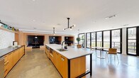 This single-floor communal space combines a well-appointed kitchen, versatile lounge areas, and dedicated meeting rooms in a cohesive, modern design. The open-plan layout is anchored by a substantial central island with a white quartz countertop, undermount sink, and warm wood cabinetry, complemented by a row of bar stools along one side. A full-length perimeter counter features matching wood cabinetry, dark countertops, stainless steel appliances, and a striking terrazzo backsplash topped with an open display shelf. Polished concrete floors extend throughout, lending an industrial yet refined character to the space. Recessed lighting and black pendant fixtures, including a sculptural multi-arm chandelier above the island, provide layered illumination. Along one wall, a series of built-in booth-style seating nooks with wood surrounds and dark upholstery offer semi-private areas for informal gatherings. Floor-to-ceiling black-framed glass doors and windows flood the interior with natural light and provide direct access to an outdoor balcony with surrounding views. A dedicated meeting area features a glass-walled conference room with a round table and task chairs, alongside additional enclosed offices accessible via wood-framed doors, ensuring the space accommodates both collaborative and private functions.