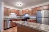 This single-floor, 2-bedroom, 1-bathroom residence offers a thoughtfully organized layout designed for comfortable everyday living. The interior features a mix of warm hardwood-style flooring in the main living and kitchen areas, transitioning to plush carpet in the bedrooms and living room.

The kitchen is well-appointed with rich wood cabinetry, granite-style countertops, stainless steel appliances including a refrigerator, electric range, and dishwasher, along with a stainless steel sink. It connects conveniently to the adjacent dining area, which features a ceiling fan and hardwood-style flooring.

The living room is bright and open, illuminated by natural light from a sliding glass door that opens to an outdoor balcony area, fitted with vertical blinds. A built-in shelving unit with a glass transom panel serves as a distinctive architectural divider between spaces.

The primary bedroom offers a comfortable footprint with carpeted flooring, a window with outdoor views, and built-in shelving. The second bedroom, also carpeted, includes a ceiling fan and window, providing a cozy and functional retreat.

The bathroom features a wide vanity with warm wood cabinetry, a long white countertop, an extended mirror, and elegant multi-globe lighting. A dedicated closet completes the layout, offering practical storage solutions throughout this well-designed residence.